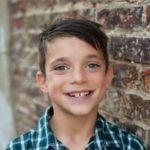 A photo of a young boy with brown hair and a plaid shirt smiling